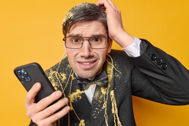 Disappointed displeased young man grabs head feels nervous feels nervous feels unhappy holds mobile phone dressed formally reacts on received unexpected message isolated over yellow background