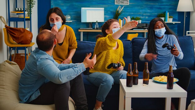 Disappointed diverse people loosing playing video games at home respecting social distancing because of corona outbreak wearing face mask against spreading virus. New normal party social distance