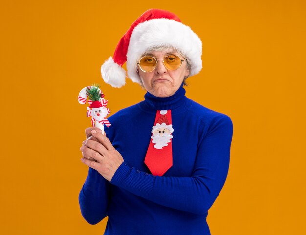 Disappointed elderly woman in sun glasses with santa hat and santa tie holding candy cane isolated on orange wall with copy space