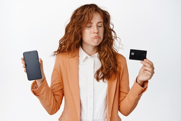 실망한 여직원이 휴대폰 화면을 보여주고 흰색 배경에 양복 차림으로 서 있는 화난 신용카드를 보고 슬픈 표정을 짓고 있다