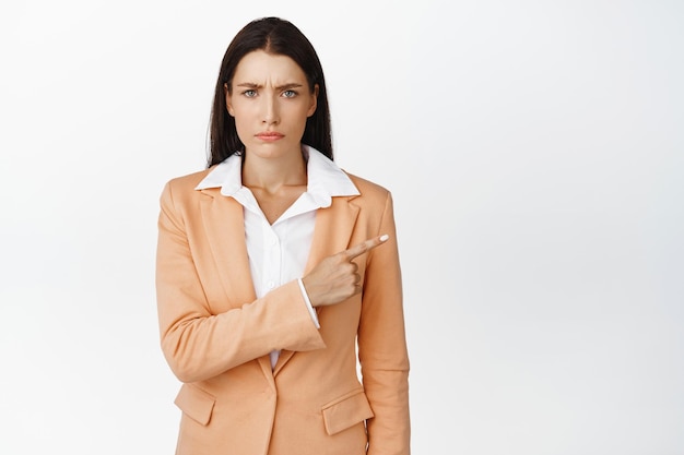 Disappointed female office worker pointing finger right entrepreneur showing logo aside and frowning displeased standing in suit over white background