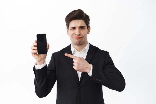 Disappointed frowning business man pointing at empty phone screen, judging bad promo, dislike website, lame promo deal, standing over white wall