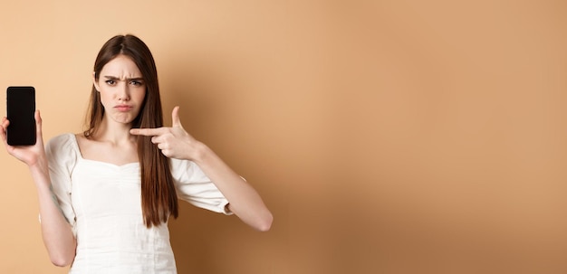 Disappointed frowning girl pointing at empty phone screen complaining at online news standing on bei