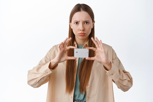 Disappointed frowning girl sulking, complaining on bank, showing credit card with displeased unfair face, being jealous, standing over white wall