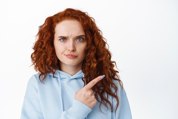 Disappointed frowning redhead woman pointing finger right at something bad, scolding or complaining, look upset and dissatisfied, white wall