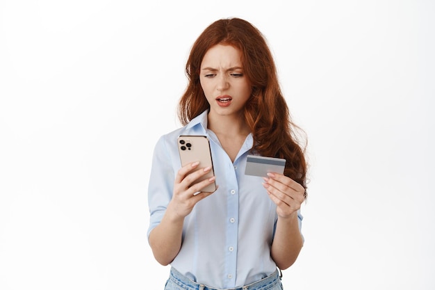 Disappointed and frustrated redhead woman looking at her phone, holding credit card, failed to pay online, having trouble with money app, standing upset against white background