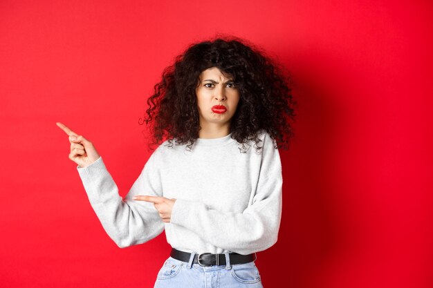 Disappointed funny woman, frowning and complaining, pointing fingers left at empty space and arguing, looking displeased, red background