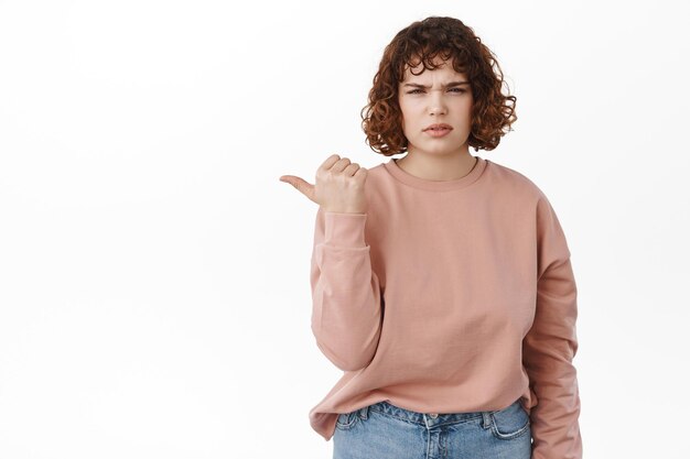 Disappointed girl complains, points left at bad promo offer, displeased by logo, dislike something and indicates at it, standing upset with frowned face against white background.