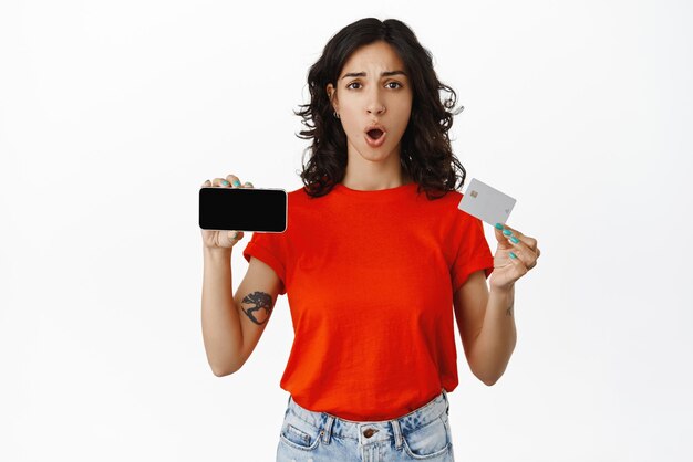 Disappointed girl customer shows horizontal phone screen and credit card looking concerned and upset with shopping experience standing over white background
