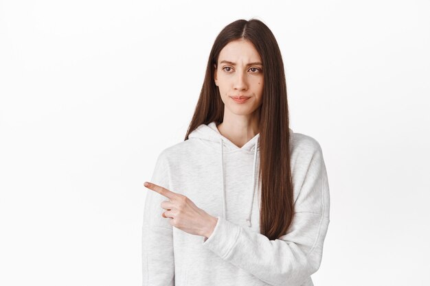 Disappointed girl grimacing, looking at something stupid or lame, judging bad promotional text, pointing aside at left copy space, dislike grimace, standing over white wall