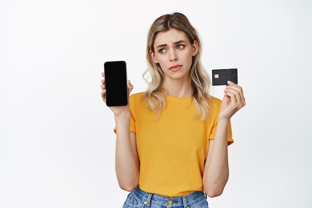 Disappointed girl shows empty smartphone and credit card sulking sad standing against white background