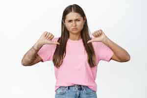 Foto gratuita ragazza delusa che strizza gli occhi e fa smorfie di antipatia, indicando se stessa, in piedi in maglietta rosa e jeans su bianco