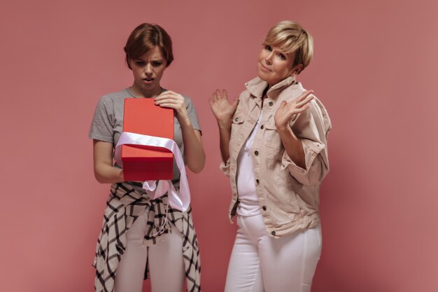 Foto gratuita ragazza delusa con i capelli castani che esamina il contenitore di regalo rosso e posa con la signora bionda in vestiti bianchi e beige su fondo rosa.