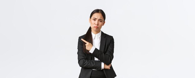 Разочарованная мрачная азиатская женщина-предприниматель теряет неудачную работу, стоя в костюме, надувая губы и указывая пальцем на неудачу Расстроенная деловая женщина делится плохими новостями на белом фоне