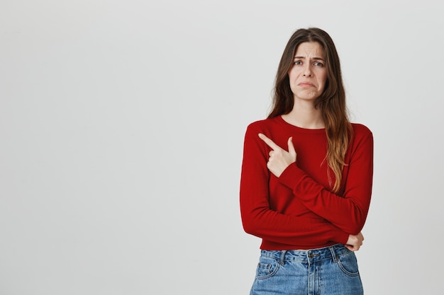 Ragazza carina cupa delusa che punta il dito nell'angolo in alto a sinistra, facendo una smorfia