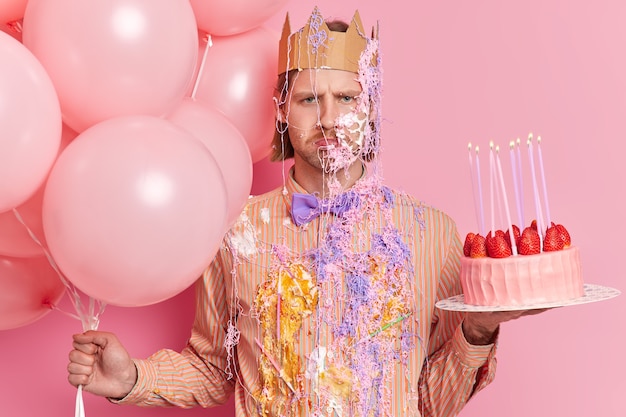 Free photo disappointed gloomy upset man smeared with cream and serpentine spray ready to celebrate birthday poses with airballoons and strawberry cake