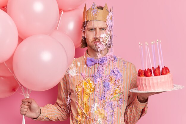 Разочарованный мрачный расстроенный мужчина, намазанный кремом и серпантином, готовый отпраздновать день рождения, позирует с воздушными шарами и клубничным тортом
