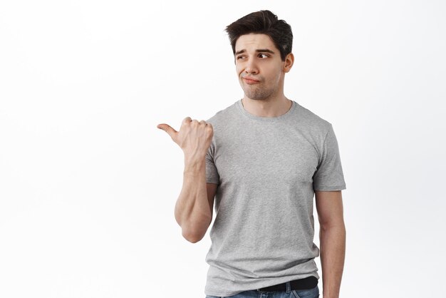 Disappointed and gloomy young man pointing left at bad product being displeased and skeptical standing unamused against white background