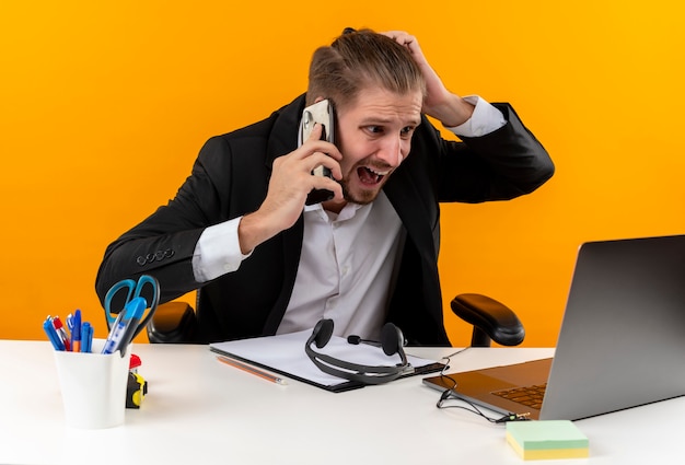 Disappointed handsome businessman in suit working on laptop talking on mobile phone looking confused and displeased sitting at the table in offise over orange background