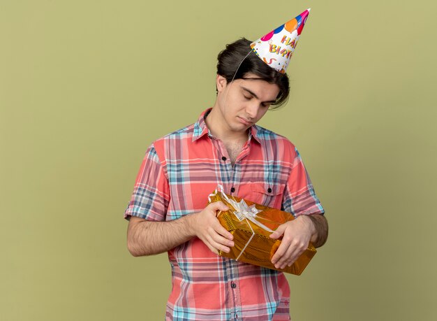 Disappointed handsome caucasian man wearing birthday cap stands with closed eyes holding gift box 