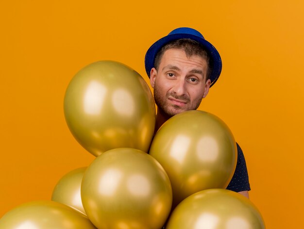 Disappointed handsome man wearing blue party hat holds helium balloons isolated on orange wall