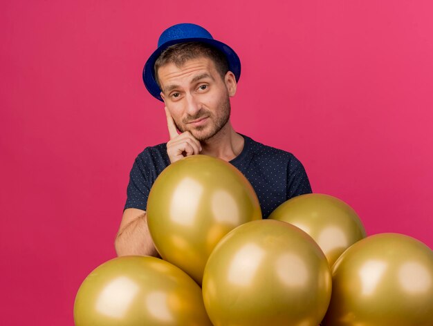 Disappointed handsome man wearing blue party hat holds helium balloons isolated on pink wall with copy space