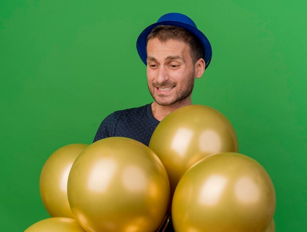 Disappointed handsome man wearing blue party hat holds and looks at helium balloons isolated on green wall with copy space