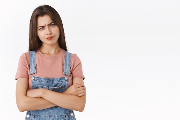 失望した、躊躇し、懐疑的な真面目な若い偉そうな女性は、Tシャツ、クロスハンドの胸、目を細めて疑わしくて笑い、嫌いな白い背景で判断を見つめています。