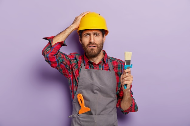 Бесплатное фото Разочарованный промышленный рабочий, одетый в защитную каску, повседневную форму, держит кисть для рисования, будучи профессиональным художником, с недовольным выражением лица, изолирован на фиолетовой стене