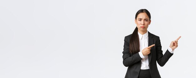 Disappointed or jealous female asian entrepreneur in suit complaining pouting and pointing fingers right Displeased saleswoman arguing look offended or sad standing white background