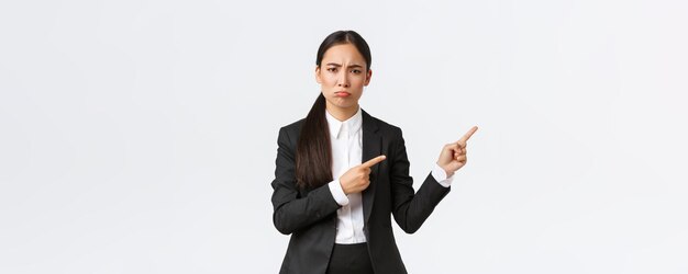 スーツを着た失望した、または嫉妬深い女性のアジアの起業家が、ふくれっ面と指を右に向けていると不平を言っている