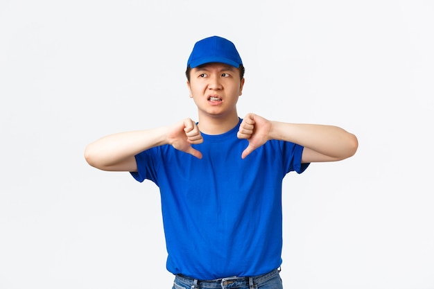 Disappointed judgemental asian male courier leave negative feedback about previous company. Delivery guy looking bothered and displeased, showing thumbs-down, standing white background.