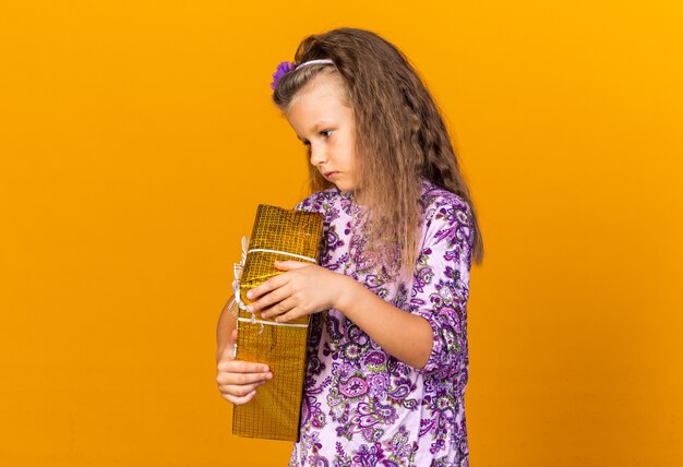 disappointed little blonde girl holding gift box and looking at side isolated on orange wall with copy space