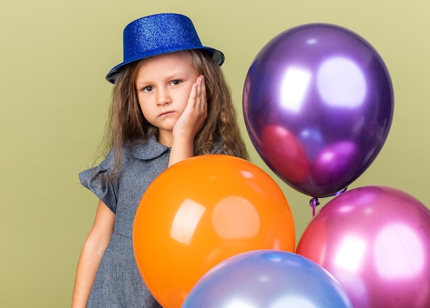 블루 파티 모자 얼굴에 손을 넣고 복사 공간 올리브 녹색 벽에 고립 된 헬륨 풍선을 들고 실망 된 작은 금발 소녀