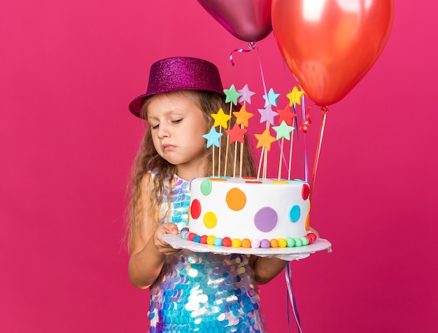 disappointed little blonde girl with purple party hat holding helium balloons and looking at birthday cake isolated on pink wall with copy space