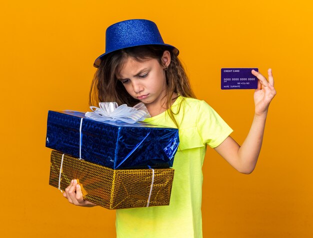 disappointed little caucasian girl with blue party hat holding gift boxes and credit card isolated on orange wall with copy space