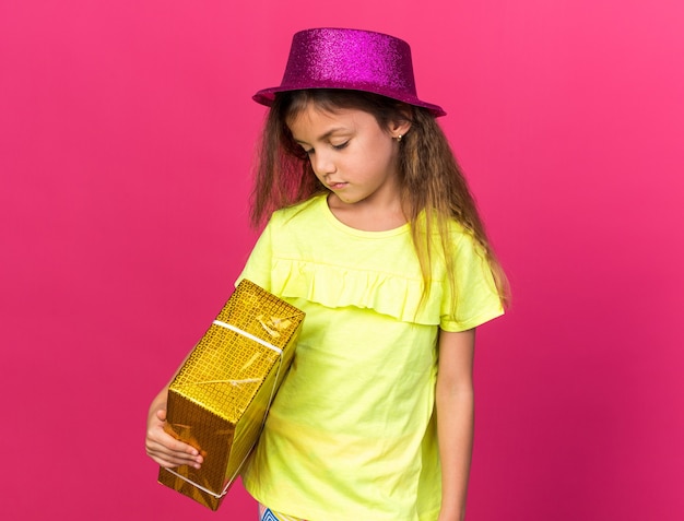 Foto gratuita delusa bambina caucasica con cappello da festa viola che tiene in mano una confezione regalo isolata sulla parete rosa con spazio per le copie