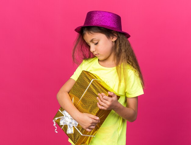 disappointed little caucasian girl with purple party hat holding and looking at gift box isolated on pink wall with copy space