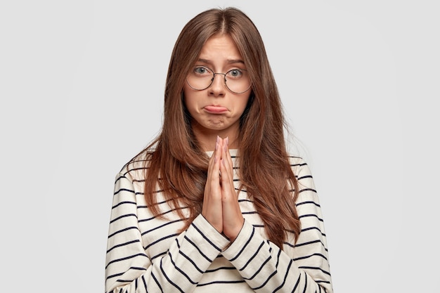 Disappointed lovely female pouts lips, has hopeful miserable expression, beggs for mercy, asks for forgiveness, dressed in casual striped sweater, isolated over white wall. Please, forgive me!