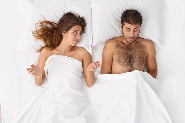 Разочарованный мужчина имеет эректильную дисфункцию во время секса, его партнерша лежит под белым одеялом, озадаченная импотенцией мужа, разводит руки в стороны. Сексуальные проблемы. Концепция здоровья мужчин
