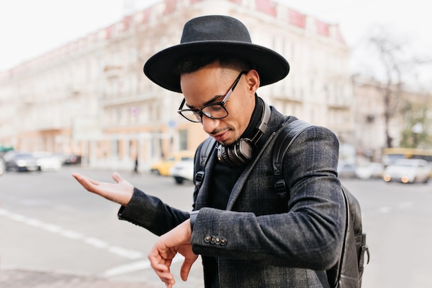 Disappointed man looking at wristwatch while waiting someone. Worried african guy in hat standing on the street.