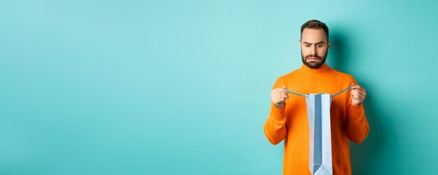 Free photo disappointed man open shopping bag and dislike gift frowning displeased standing in orange sweater a