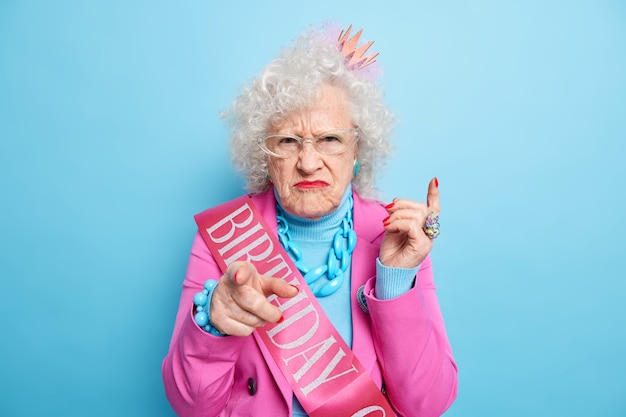 Disappointed mature woman squints face points directly at you invites on her birthday party dressed in festive clothes being angry with offensive words about her age wears bright makeup stands indoor