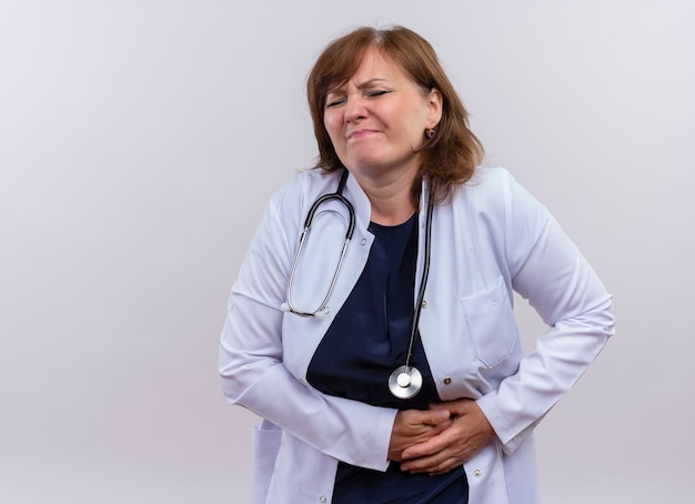Disappointed middle-aged woman doctor wearing medical robe and stethoscope hands on kidneys suffering from kidney pain on isolated white wall with copy space