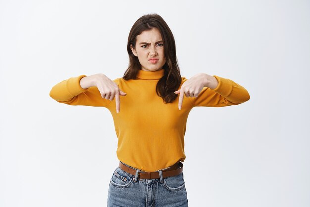 Disappointed moody girl complaining, grimacing and pointing fingers down at awful advertisement, dislike and disapprove something, white wall