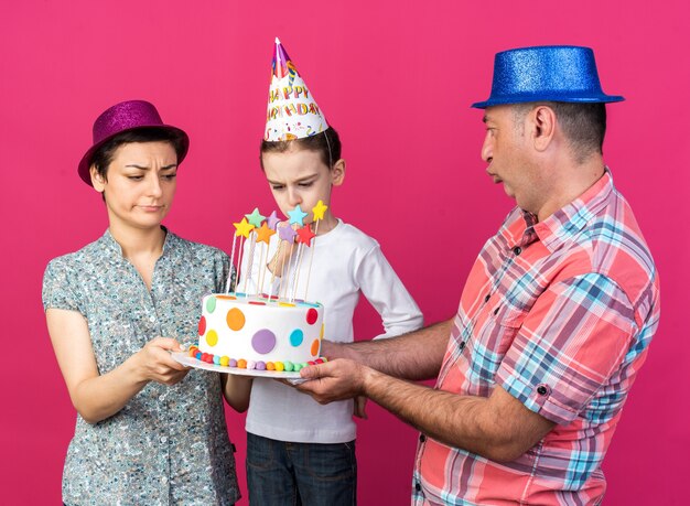 disappointed mother and father with party hats holding birthday cake together looking at their unpleased son isolated on pink wall with copy space