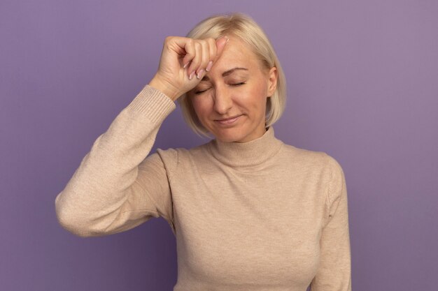 Free photo disappointed pretty blonde slavic woman puts hand on forehead standing with closed eyes on purple