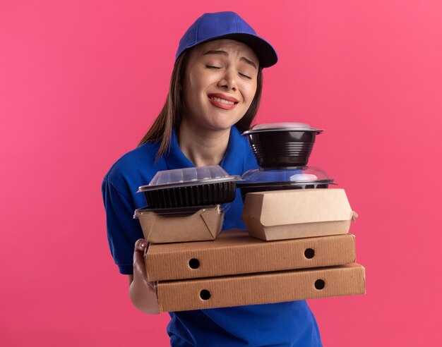 Disappointed pretty delivery woman in uniform holds food package and containers on pizza boxes