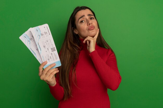 Disappointed pretty woman puts hand on chin holds air tickets isolated on green wall