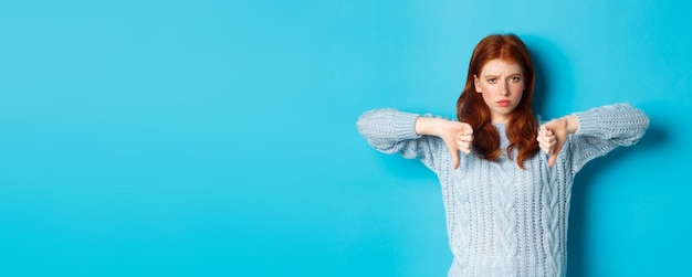 Бесплатное фото Разочарованная рыжеволосая девушка в свитере показывает большой палец вниз, осуждая плохой продукт, не соглашается и не любит профессионала